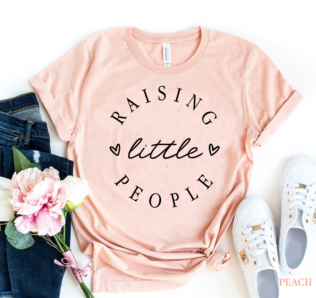 Pink t-shirt with 'RAISING little PEOPLE' text on a white background with jeans, sneakers, and flowers.